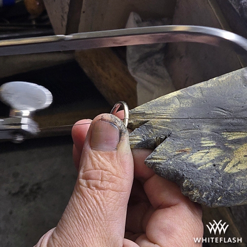 A jeweler carefully sawing through a ring on a workbench using a precision jeweler's saw, preparing the piece for resizing. The craftsman's hands, holding the ring securely against a worn bench pin, show signs of experience and meticulous craftsmanship.