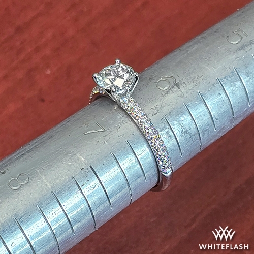 A diamond engagement ring with a round brilliant center stone set in a white goldpavé band, placed on a metal ring mandrel for sizing. The ring showcases a sparkling diamond with a secure four-prong setting and a delicate, shimmering band adorned with small accent diamonds.